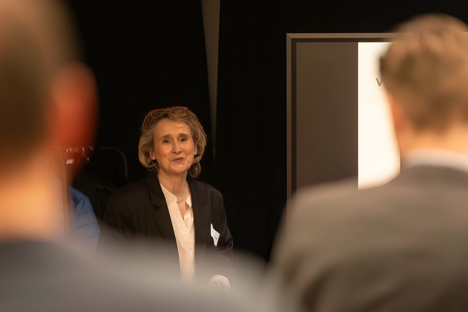 Christiane Schicker spricht frontal zu einer Gruppe von Menschen. In der Nahaufnahme erkennt man ihr Gesicht, sie bewegt lächelnd mit aufmerksamen Blick den Mund und spricht frontal zum Publikum. Eine Lesebrille ist locker über der Stirn in den Haaren gesteckt. Ihr Gesicht vor dunklem Hintergrund wird im Vordergrund gerahmt und fokussiert von den unscharfen Silhouetten von zwei Personen im Publikum, die zuhören, ihren Rücken dem Fotografen zugewendet.